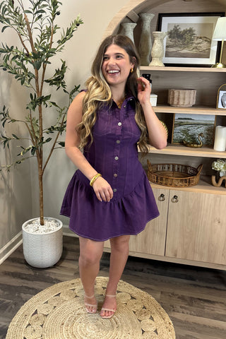 Woman in a purple dress standing in a room with a plant and wooden cabinet.