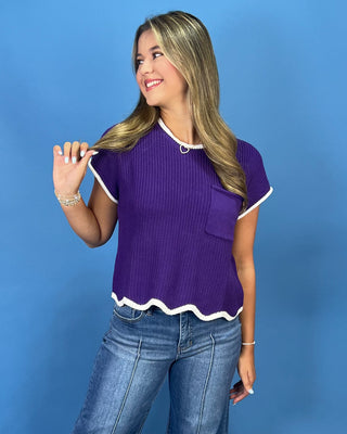 A woman modeling a purple short-sleeve ribbed knit top with white trim, paired with blue jeans, against a solid blue studio background.