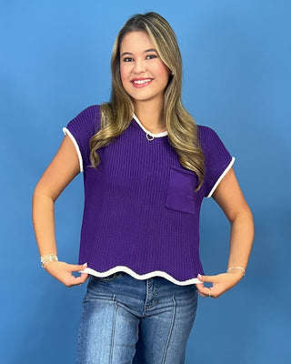 A woman modeling a purple short-sleeve ribbed knit top with white trim and a chest pocket, paired with blue jeans, against a solid blue background.