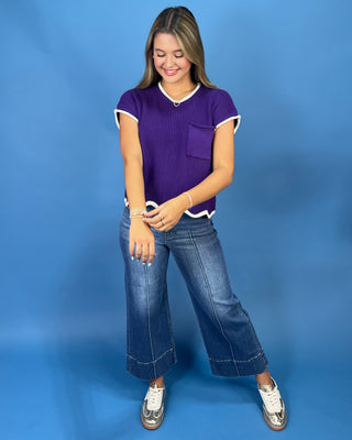 A young woman modeling a purple short-sleeve ribbed knit top with white trim, paired with wide-leg blue jeans and white sneakers, standing against a solid blue studio backdrop.