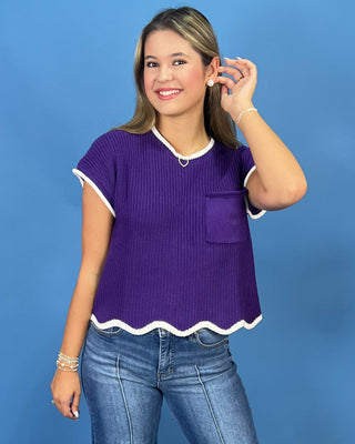 A woman modeling a purple short-sleeve ribbed knit top with white trim, a scalloped hem, and a chest pocket, paired with blue jeans against a solid blue background.