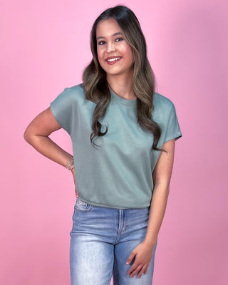 Muted sage green short sleeve top with a relaxed fit and soft, lightweight fabric, paired with cropped wide leg denim jeans and neutral wedge sandals, styled against a pink backdrop.