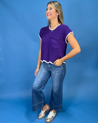 A woman modeling a purple short-sleeve ribbed knit top with white trim and a chest pocket, paired with blue jeans and white sneakers, against a solid blue studio backdrop.