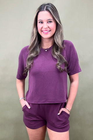 A woman wearing a matching plum-colored lounge set with a short-sleeve top and drawstring shorts, paired with white sneakers, standing against a plain olive backdrop