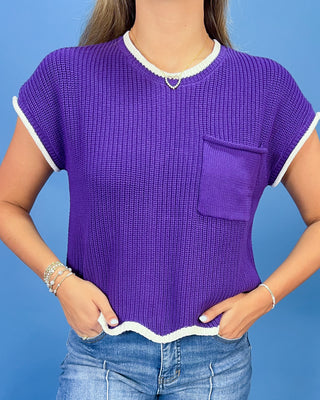 A woman wearing a purple short-sleeve ribbed knit top with white trim on the collar, sleeve cuffs, and hem, paired with blue jeans against a solid blue background.