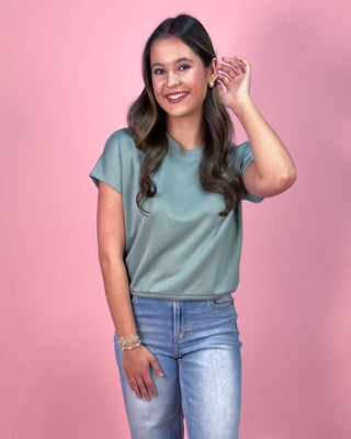 Muted sage green short sleeve top with a relaxed fit and soft, lightweight fabric, paired with cropped wide leg denim jeans and neutral wedge sandals, styled against a pink backdrop.