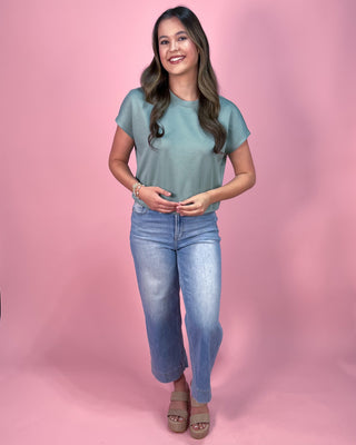 Muted sage green short sleeve top with a relaxed fit and soft, lightweight fabric, paired with cropped wide leg denim jeans and neutral wedge sandals, styled against a pink backdrop.
