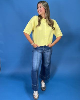 A woman modeling a pale yellow short-sleeve button-up blouse with blue jeans and white sneakers, standing against a solid blue studio backdrop.