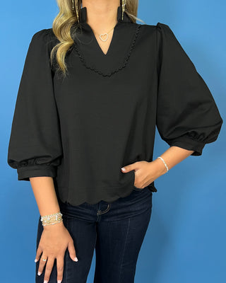 Black scalloped blouse with a split neckline, puff sleeves, scalloped trim, and a polished feminine fit.