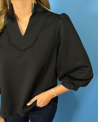 Black scalloped blouse with a split neckline, puff sleeves, scalloped trim, and a polished feminine fit.