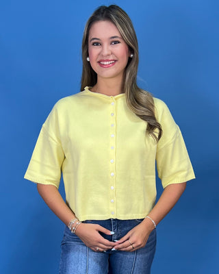 A woman modeling a pale yellow short-sleeve button-front top with a round neckline, paired with blue jeans, against a solid blue studio background.