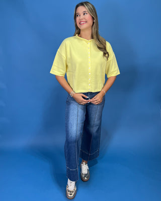 A woman modeling a pale yellow short-sleeve button-up shirt with blue jeans and metallic sneakers, standing against a solid blue studio backdrop.