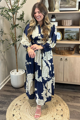 Woman in a blue and white patterned dress standing in a room with a plant and wooden cabinet.