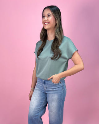 Muted sage green short sleeve top with a relaxed fit and soft, lightweight fabric, paired with cropped wide leg denim jeans and neutral wedge sandals, styled against a pink backdrop.