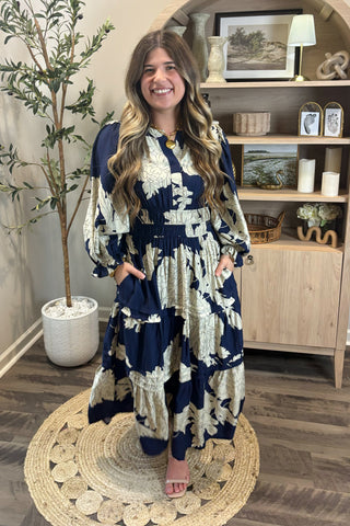 Woman in a blue and white floral dress standing in a room with wooden flooring and a plant.