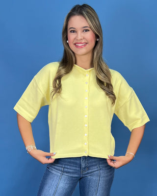 a pale yellow short-sleeve button-up knit top with white buttons, paired with blue jeans, against a solid blue studio background.