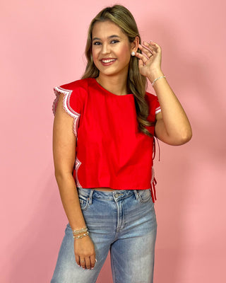 Woman wearing a red top with lace details and blue jeans against a pink background