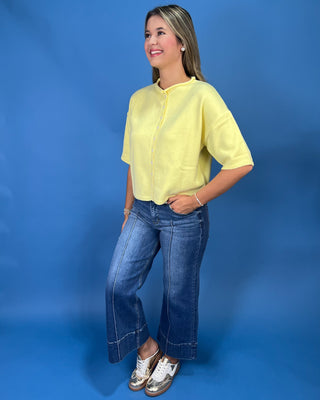 A woman modeling a pale yellow short-sleeve button-front top with blue jeans and white sneakers, standing against a solid blue studio backdrop.