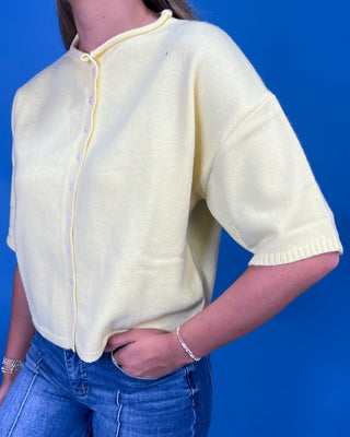 A woman wearing a pale yellow short-sleeve knit cardigan with a button placket and ribbed hem, paired with blue jeans, set against a solid blue background.