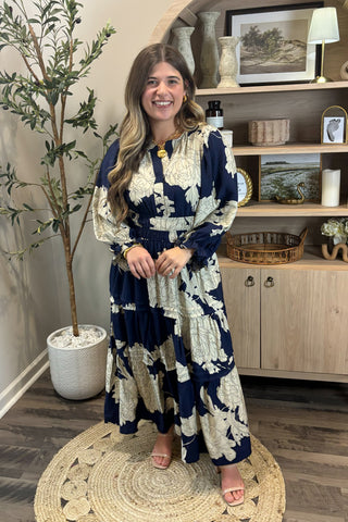Woman in a blue and white floral dress standing in a room with a plant and shelves.