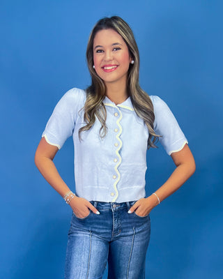 A woman modeling a light blue short-sleeve button-up blouse with a wavy front button placket and scalloped edges, paired with blue jeans, against a solid blue studio background.