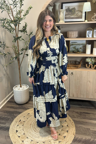 Woman wearing a blue and white floral dress standing in a room with a plant and shelves.