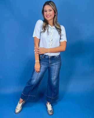 A woman posing in a light blue short-sleeve blouse with a yellow ruffle trim and blue wide-leg jeans, standing against a solid blue studio backdrop.