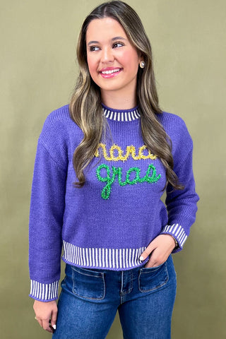 Woman wearing a purple sweater with 'Mama Gras' text, standing against a beige background