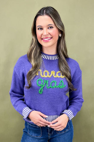 Woman wearing a purple sweater with 'Mardi Gras' text, standing against a beige background