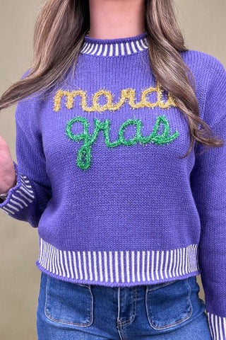 Purple sweater with 'mardi gras' text worn by a person against a beige background