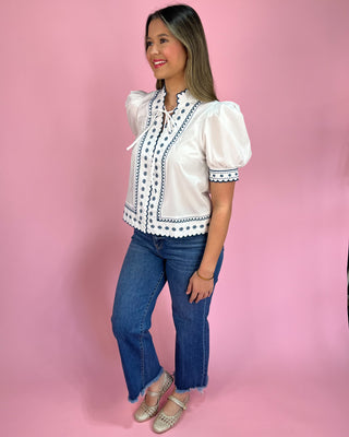 Woman wearing a white blouse with black patterns and blue jeans against a pink background