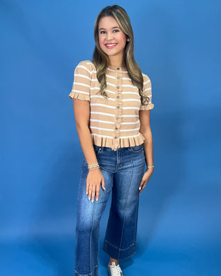 A young woman posing in a beige and white striped short-sleeve crop top with a ruffled hem, paired with blue jeans, aga