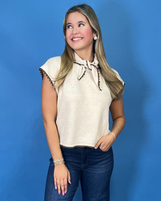 Cream sleeveless tie-neck knit top with brown contrast trim, styled with dark wash wide leg jeans.