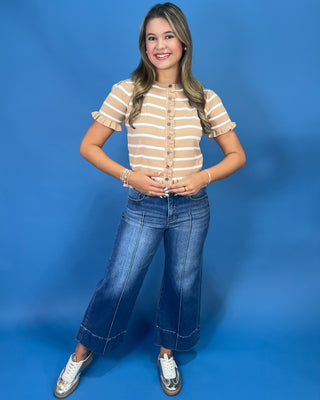 A young woman posing in a beige and white striped short-sleeve button-up top, blue wide-leg jeans, and white sneakers against