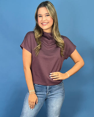 A woman modeling a short-sleeve, mock-neck top in a dusty plum purple color, paired with blue jeans, against a solid blue studio background.