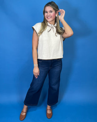 Cream sleeveless tie-neck knit top with brown contrast trim, styled with dark wash wide leg jeans.