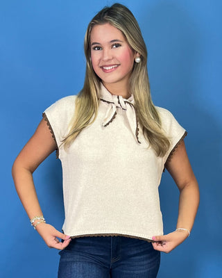 Cream sleeveless tie-neck knit top with brown contrast trim, styled with dark wash wide leg jeans.