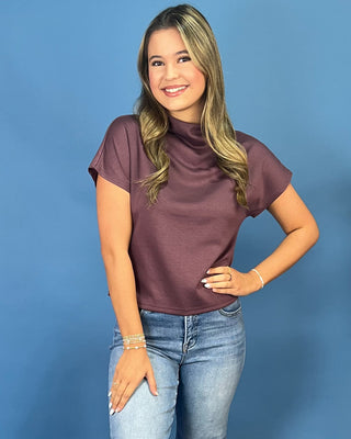 A woman modeling a short-sleeve mock neck top in a dusty plum purple color, paired with blue jeans, against a solid blue studio background.