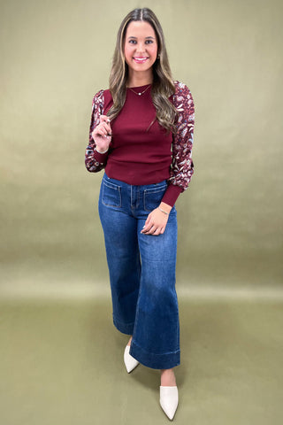 Woman wearing a burgundy top, floral cardigan, blue jeans, and white shoes on a green background