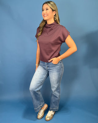 A woman modeling a short-sleeve mock neck top in a dusty plum purple color, paired with light-wash blue jeans and metallic gold loafers, standing against a solid blue studio backdrop.