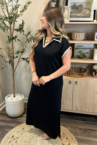 Woman in a black dress standing in a room with a plant and wooden cabinet.