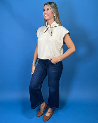 Cream sleeveless tie-neck knit top with brown contrast trim, styled with dark wash wide leg jeans.