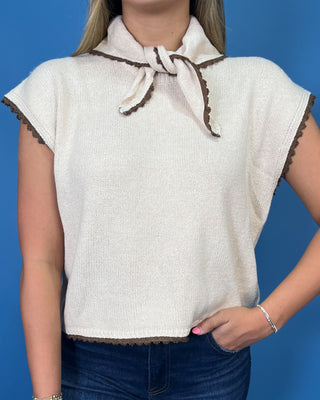 Cream sleeveless tie-neck knit top with brown contrast trim, styled with dark wash wide leg jeans.