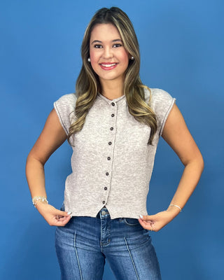 A woman modeling a light heather gray sleeveless button-front knit top with dark buttons, paired with blue jeans, against a solid blue studio background.
