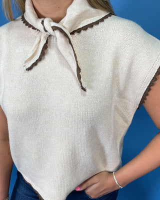 Cream sleeveless tie-neck knit top with brown contrast trim, styled with dark wash wide leg jeans.