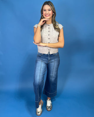 A woman posing in a light beige textured short-sleeve button-up top, blue cropped flare jeans, and white sneakers with gold trim, set against a solid blue studio backdrop.