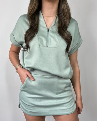 Smiling woman standing against a neutral background wearing a sage green short-sleeve zip-neck top with a matching mini skirt, styled with metallic gold sneakers and hands in pockets.