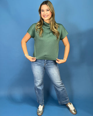 A woman modeling a sage green short-sleeve mock neck top with light-wash blue jeans and white sneakers, standing against a solid blue studio backdrop.