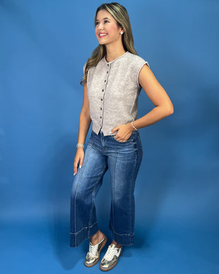 A woman modeling a light beige textured sleeveless button-front top with dark buttons, paired with wide-leg blue jeans and metallic sneakers, set against a solid blue studio background.