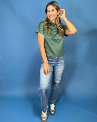 A woman modeling a short-sleeve green t-shirt and light-wash blue jeans against a solid blue studio backdrop.